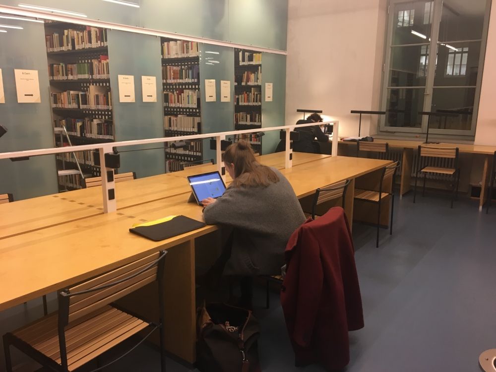 ZUG-F, Bibliothek: Blick auf die Lernplätze und Bücherregale vom Eingang her zur linken Seite. Jeder Tisch hat direkt unter der Lampe einen Stromanschluss.