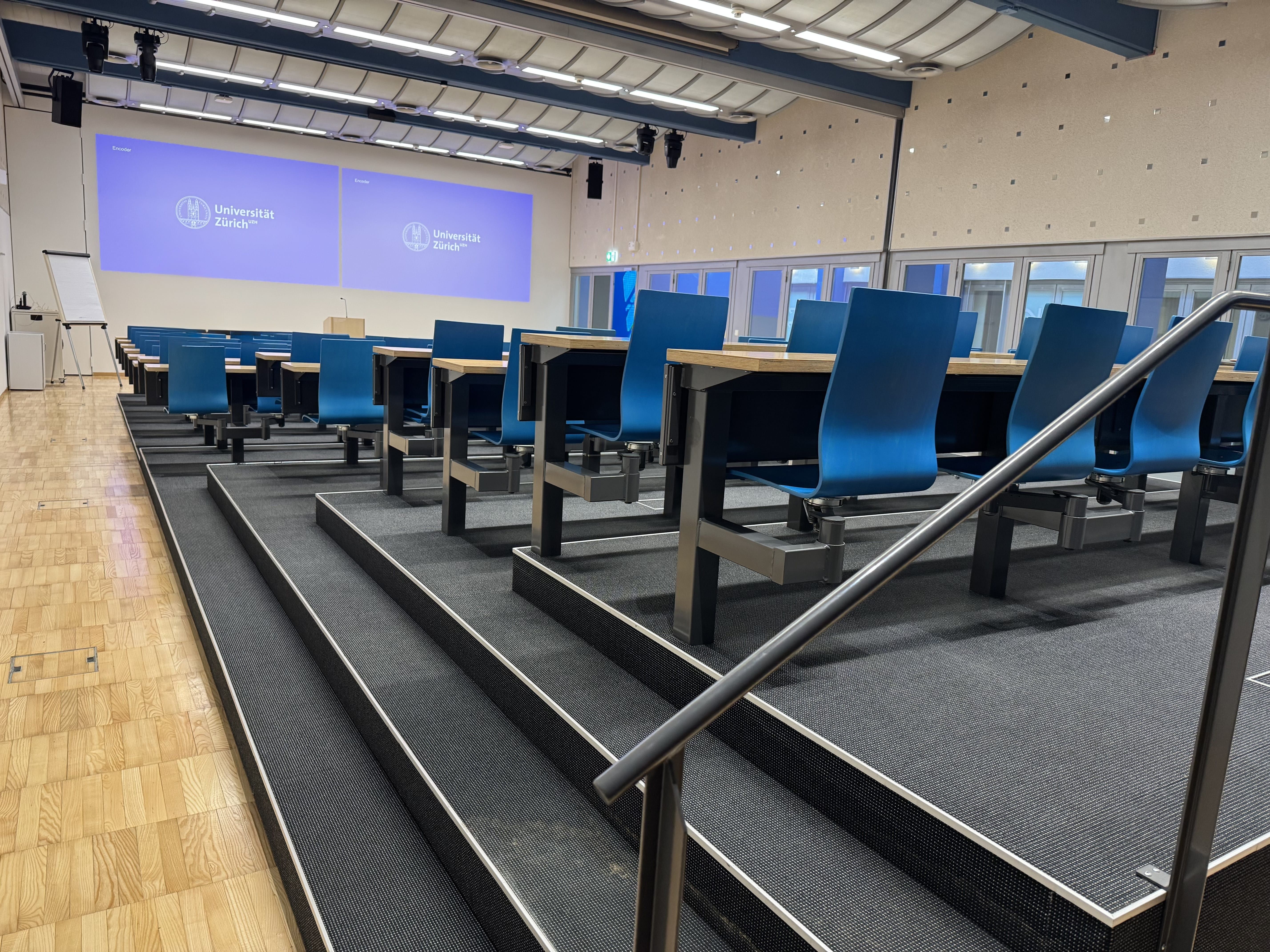 Hörsaal BIN-0-K.02: Ansicht auf den Raum zum Dozierendenpult hin. Stufen führen zu den hinteren Sitzreihen. Alle Sitzreihen können nur über eine schmale Schwelle erreicht werden, mit Ausnahme des Tisches für Rollstühle.