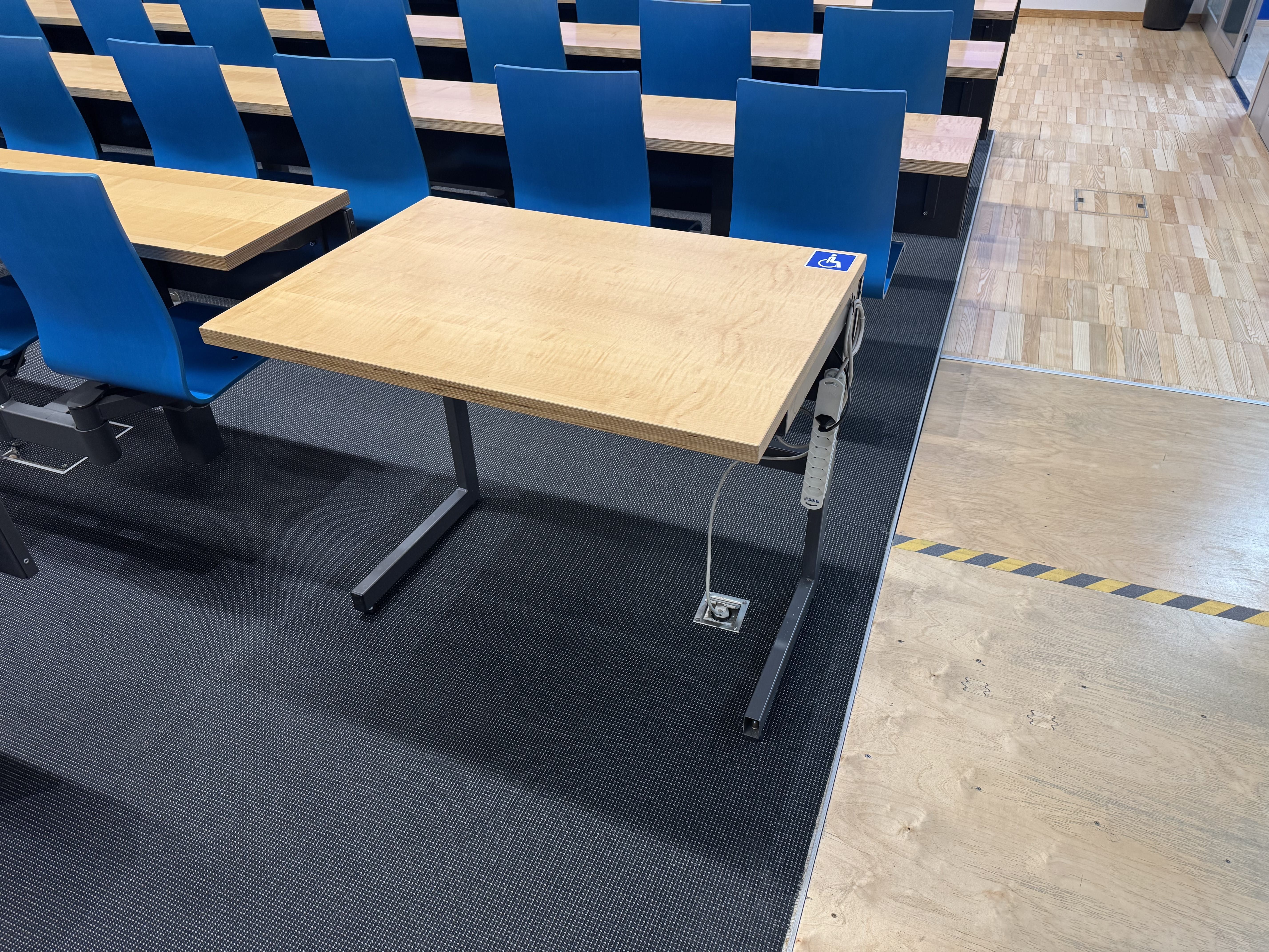 Hörsaal BIN-0-K.02: Ansicht auf den Tisch mit Rollstuhl-Signet direkt neben der Rampe und Eingangstür.
