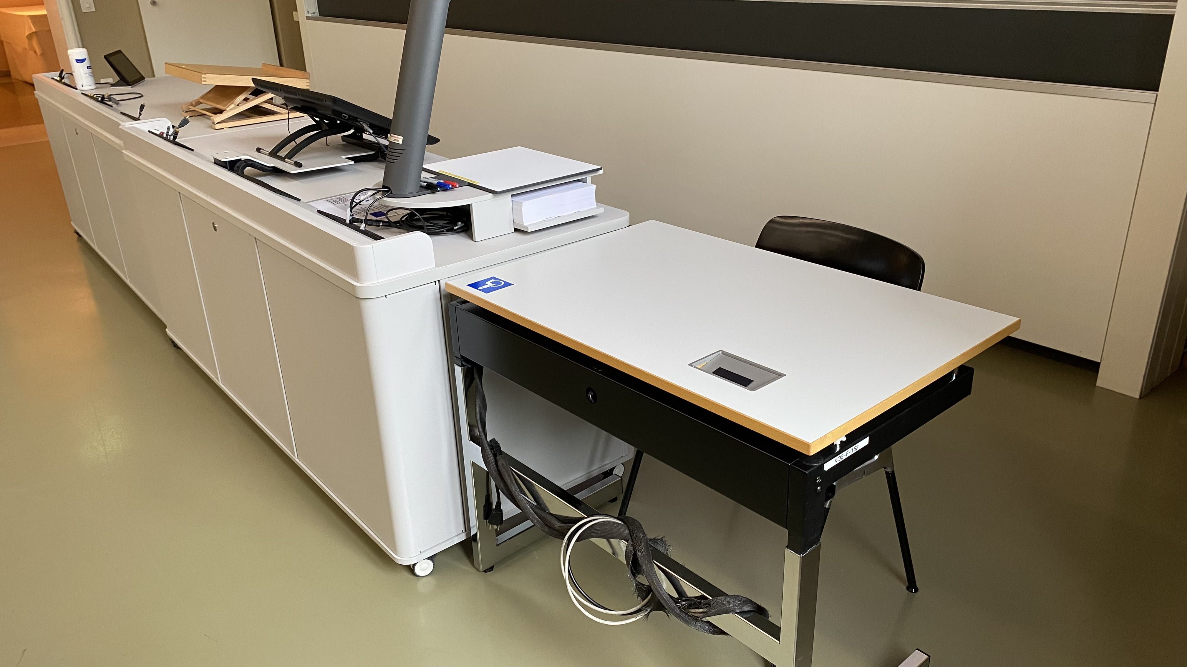 Seminarraum KO2-F-152: Ansicht auf den elektrisch höhenverstellbaren Tisch mit Rollstuhl-Signet neben dem Dozierendenpult.