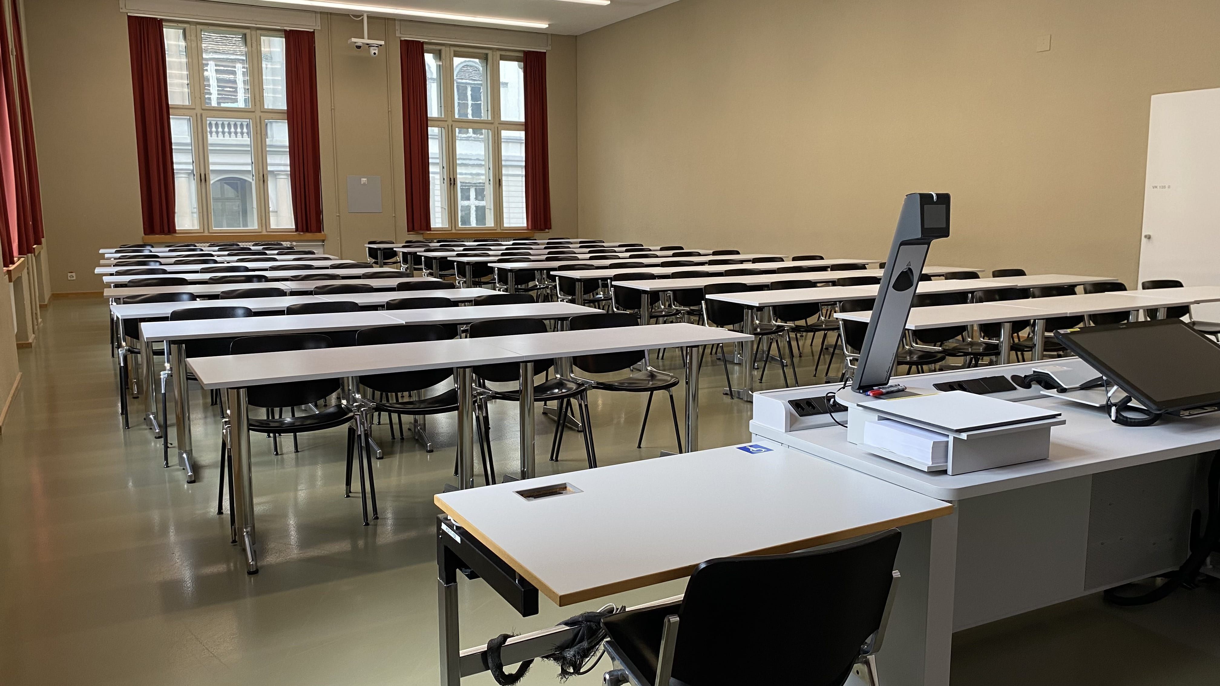 Seminarraum KO2-F-152: Ansicht auf den Raum vom Dozierendenpult aus. Direkt links neben dem Dozierendenpult befindet sich ein elektrisch höhenverstellbarer Tisch mit Rollstuhl-Signet.