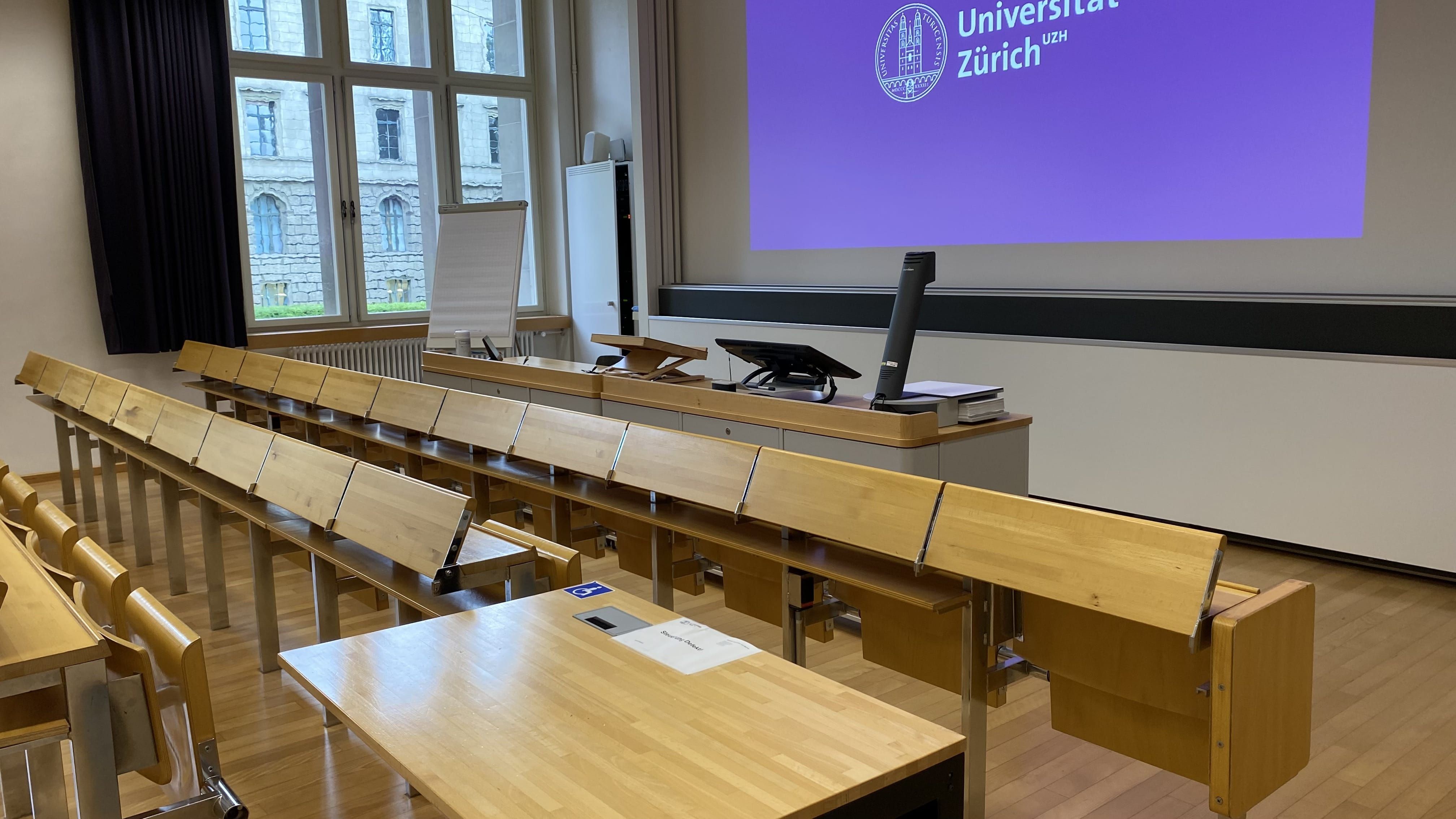 Hörsaal KO2-F-150: Ansicht auf den elektrisch höhenverstellbaren Tisch mit Rollstuhl-Signet. Der Tisch befindet sich in der zweiten Sitzreihe nahe der Eingangstüre.