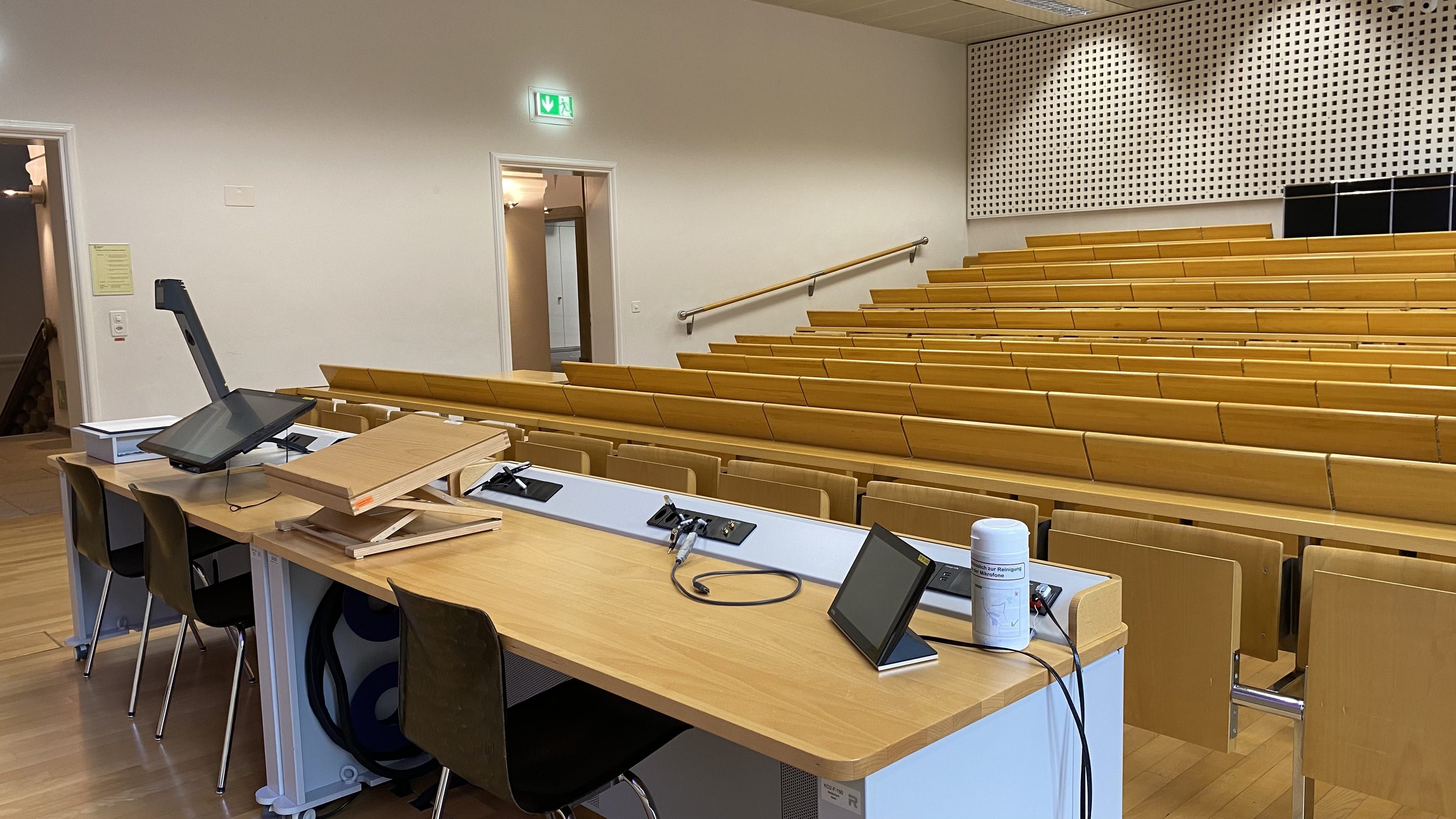 Hörsaal KO2-F-150: Ansicht auf das Dozierendenpult. Beide Eingänge in den Raum befinden sich auf der linken Seite des Bildes.