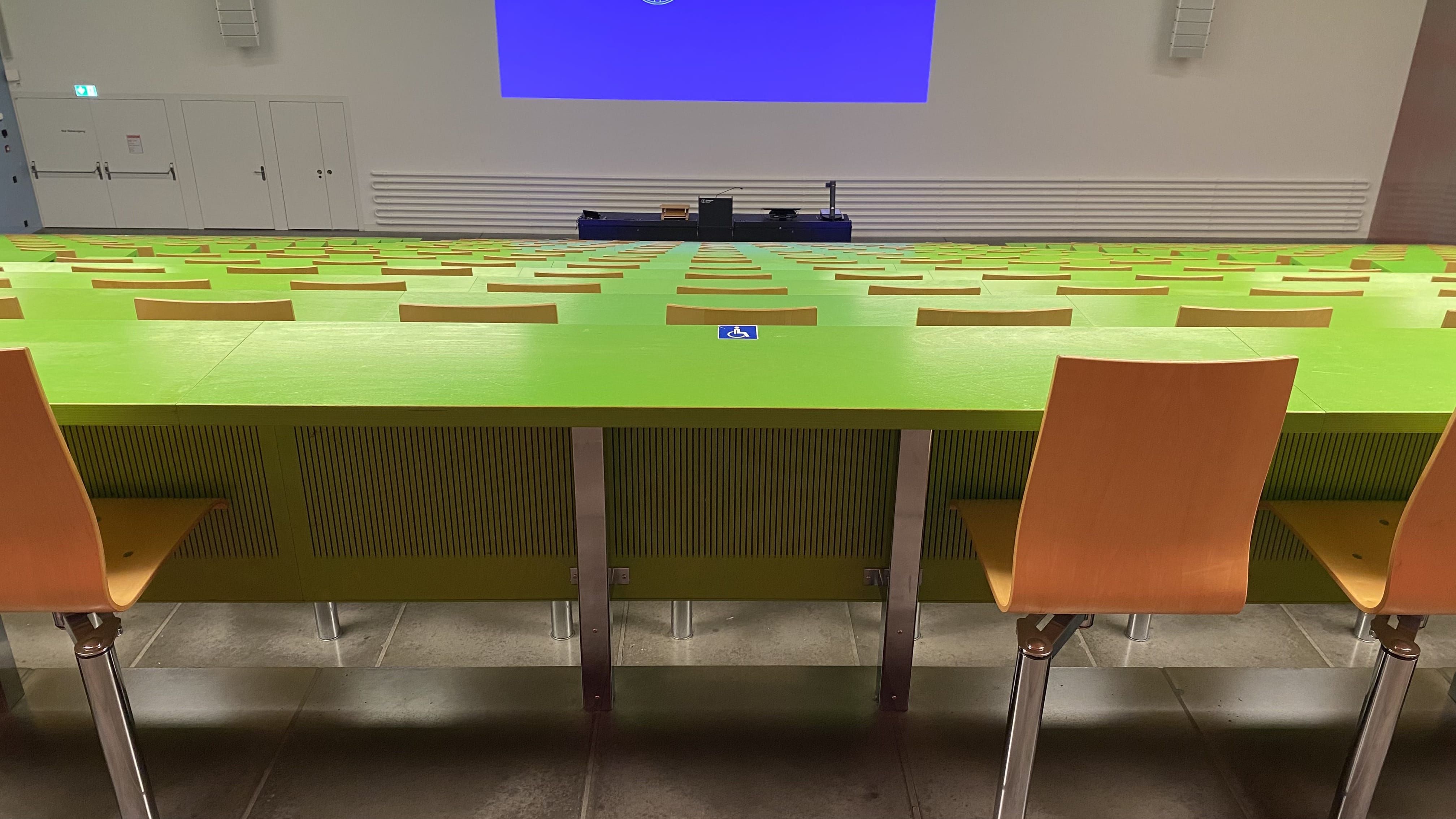 Hörsaal KOH-B-10: Ansicht auf den zweiten Tisch mit Rollstuhl-Signet ohne fixe Bestuhlung. Dieser Tisch befindet sich in der Mitte des mittleren Segments der hintersten Reihe.