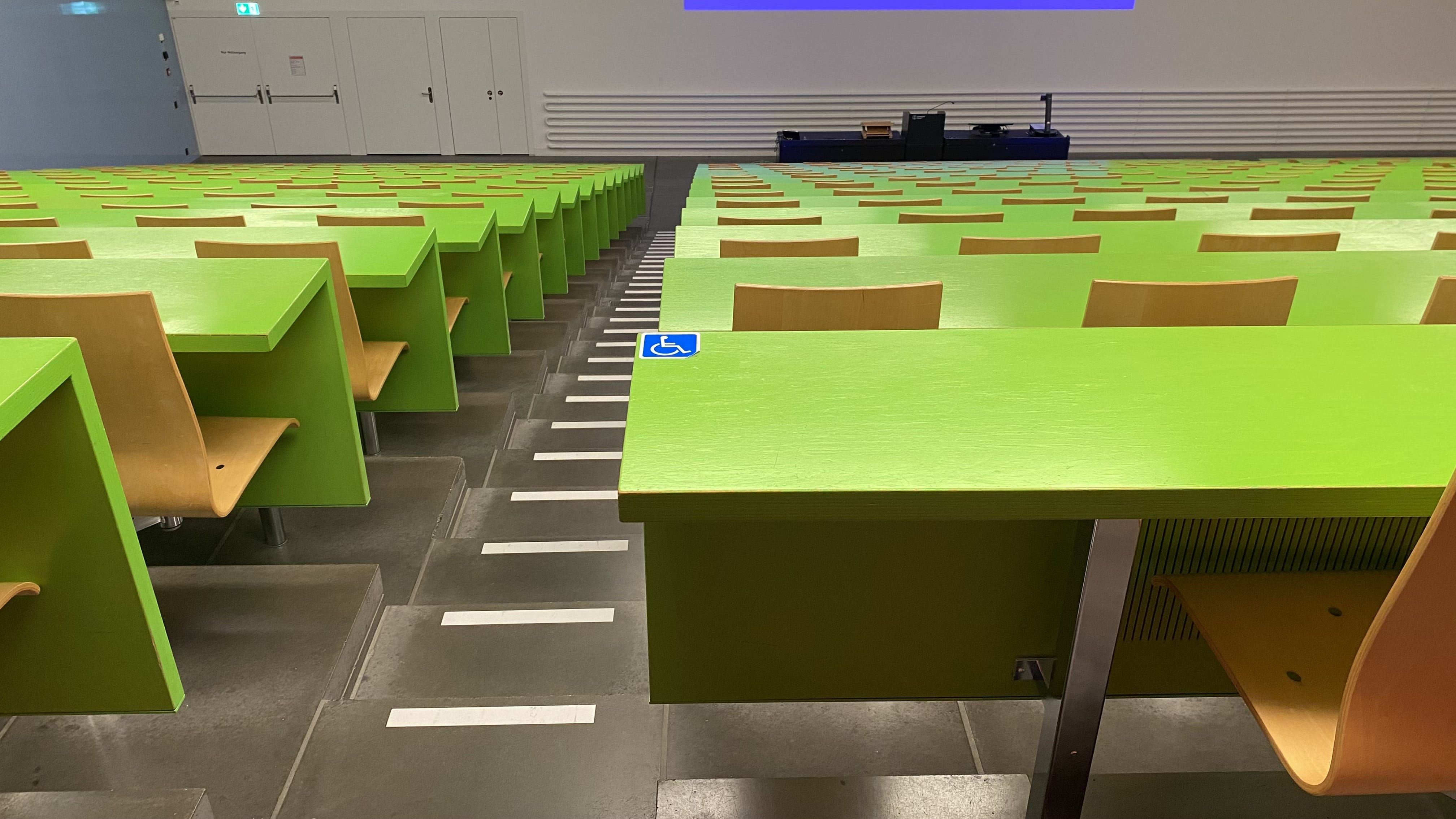Hörsaal KOH-B-10: Ansicht auf den ersten von drei Tischen mit Rollstuhl-Signet und keiner fixen Bestuhlung. Dieser Tisch befindet sich ganz links im mittleren Segment der hintersten Reihe.