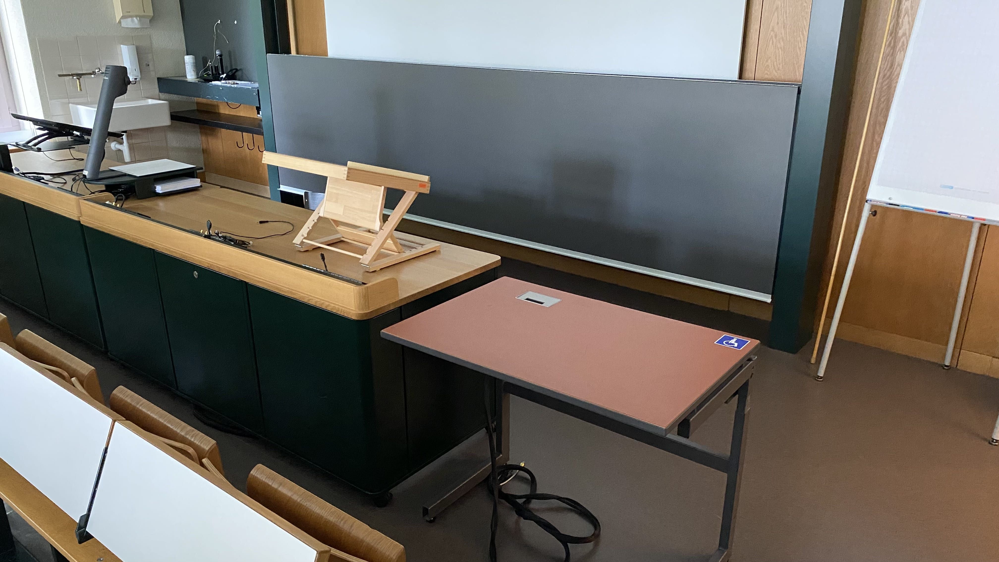 Hörsaal RAK-E-8: Ansicht auf den Tisch mit Rollstuhl-Signet direkt neben dem Dozierendenpult und nahe der Eingangstür.