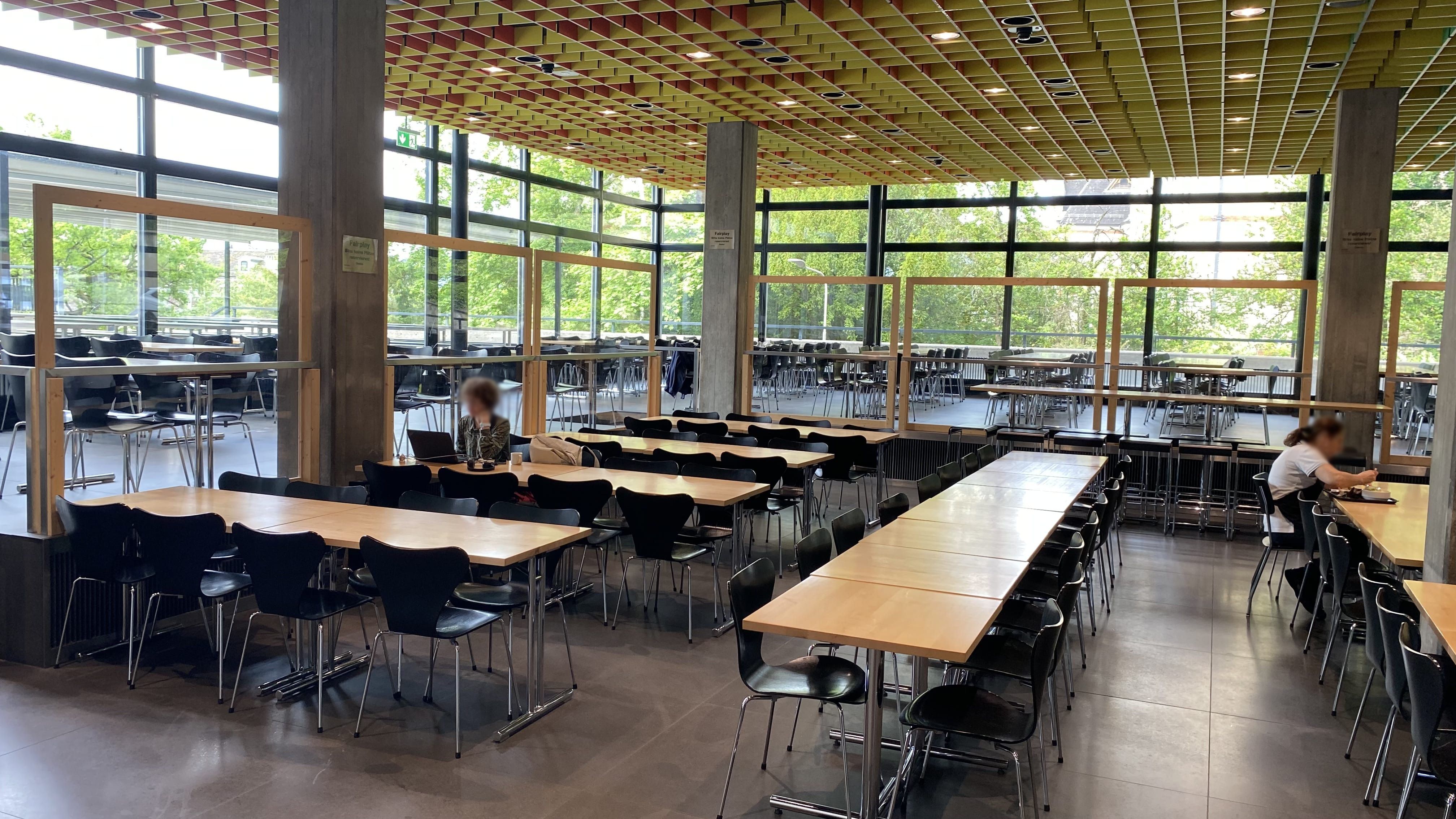 MEN-A, Mensa: Ansicht auf die ebenerdigen Tische von der Eingangstür aus. Die Mensa verfügt über einen ebenerdigen Sitzbereich, und einen erhöhten Sitzbereich, der nur über Stufen erreichbar ist.