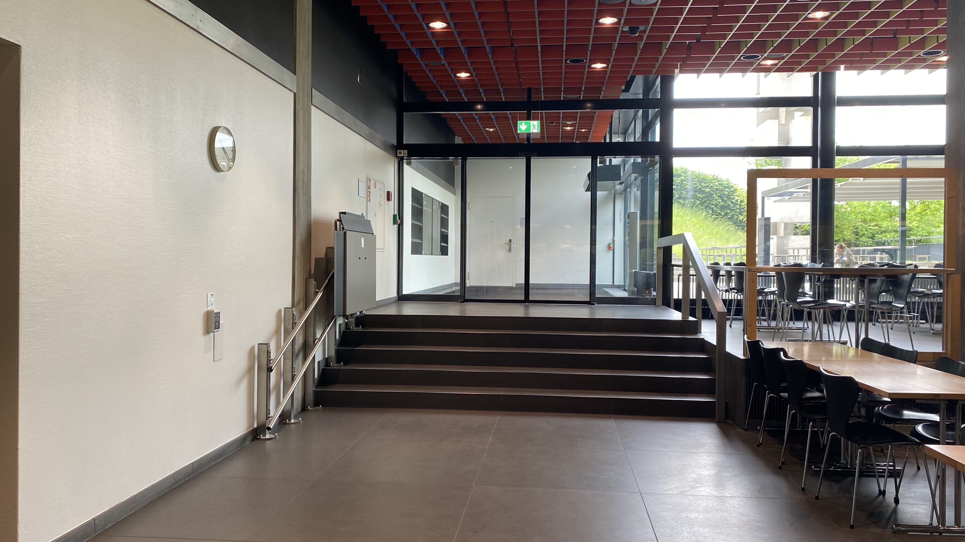 MEN-A, Mensa: Ansicht auf die Stufen links von der Eingangstür, die zum Aussenbereich der Mensa führen. Es gibt einen Treppenlift.