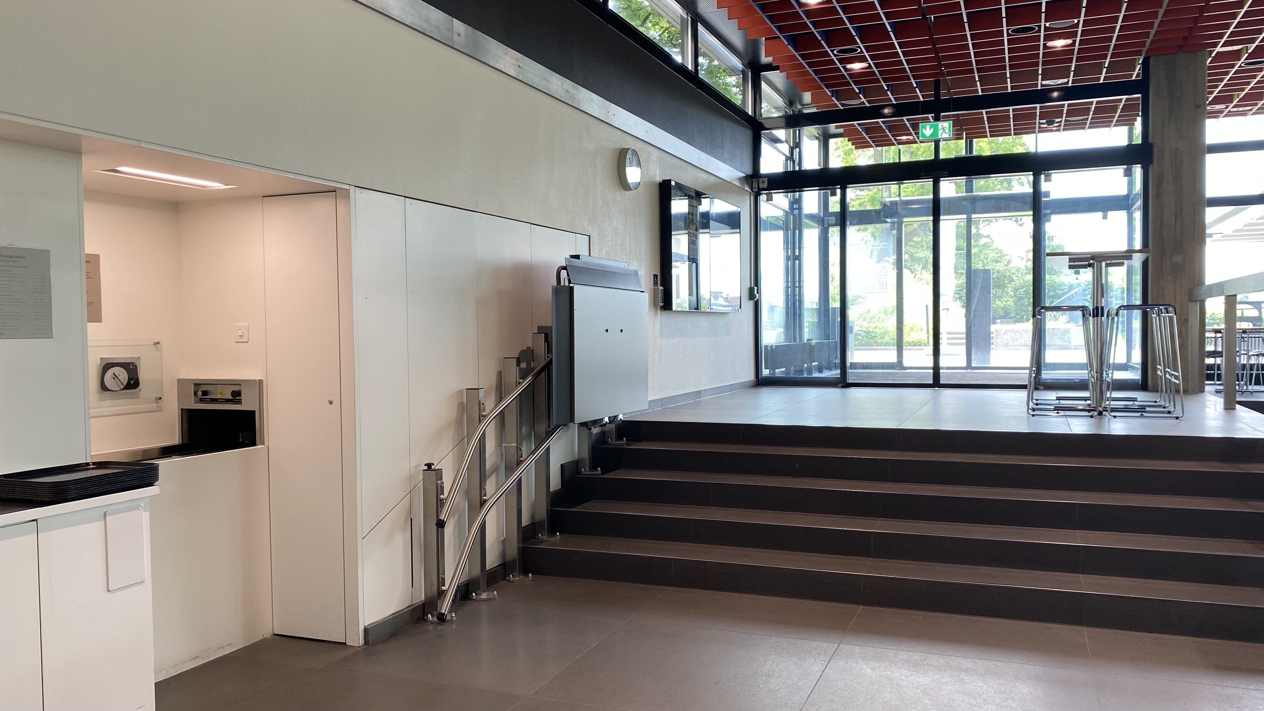 MEN-B, Mensa: Ansicht auf die Stufen links von der Eingangstür, die zum Aussenbereich der Mensa führen. Es gibt einen Treppenlift.