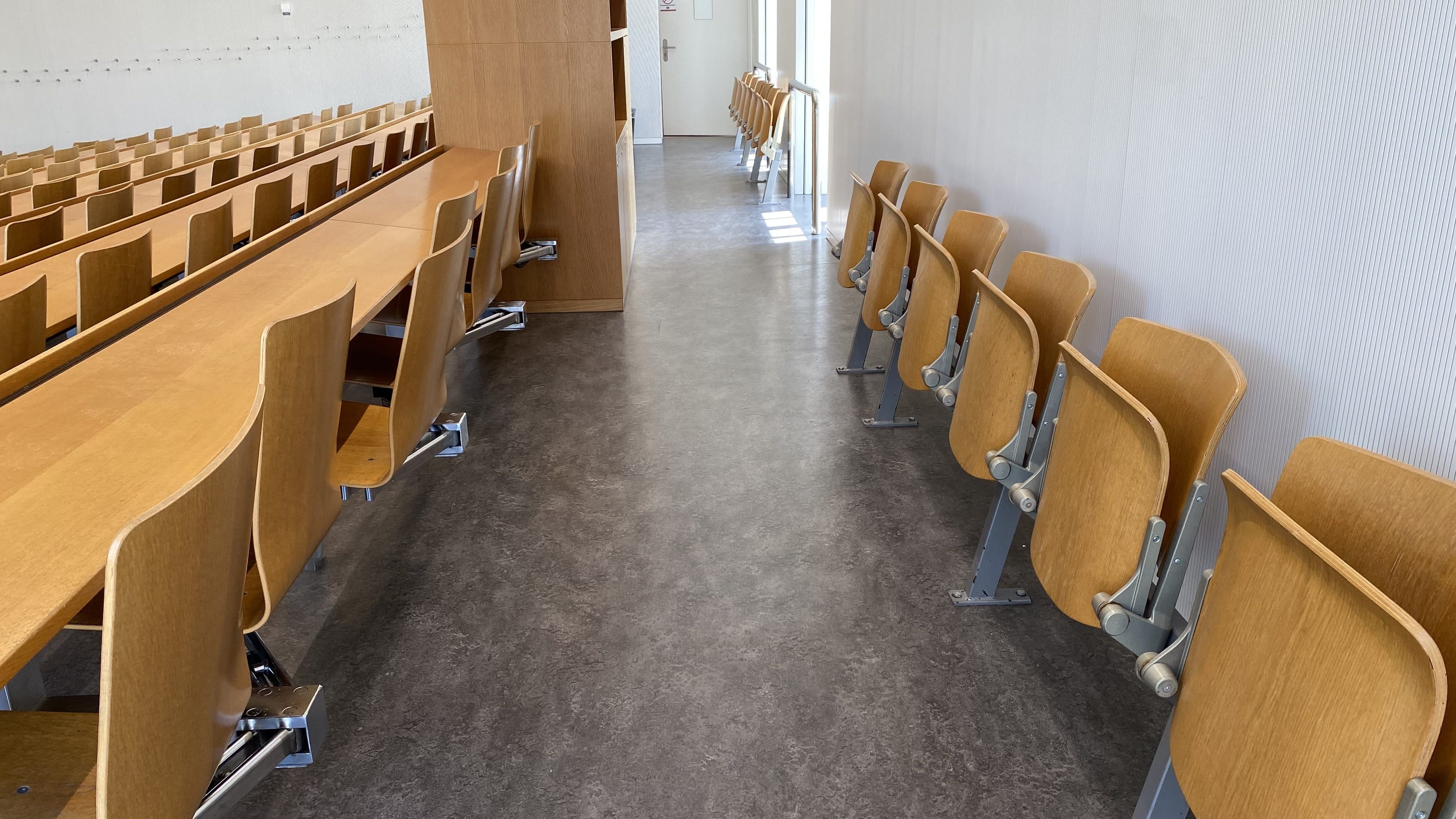 Seminarraum RAI-G-41: Ansicht auf die hinterste Reihe des Raumes. Der Durchgang zwischen Sitzmöglichkeiten mit und ohne Tisch ist breit. Auf dieser Ebene gibt es eine zweite Eingangstüre, im Bild in der Mitte ersichtlich.