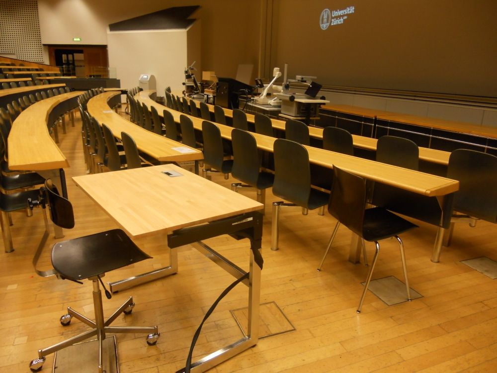 Hörsaal KO2-F-180: Elektrisch höhenverstellbares Pult in der dritten Reihe auf der rechten Seite und unmittelbar davor in der zweiten Reihe der für Rollstuhlfahrende reservierte normale Tische.