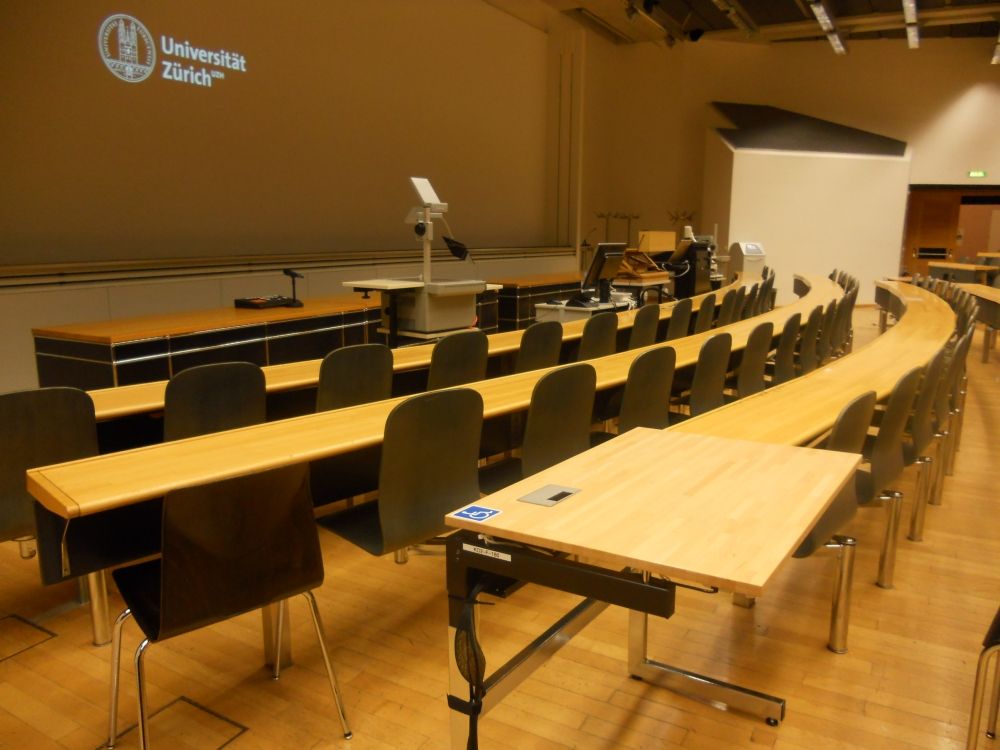 Hörsaal KO2-F-180: Elektrisch höhenverstellbares Pult in der dritten Reihe auf der linken Seite und unmittelbar davor in der zweiten Reihe der für Rollstuhlfahrende reservierte normale Tische.