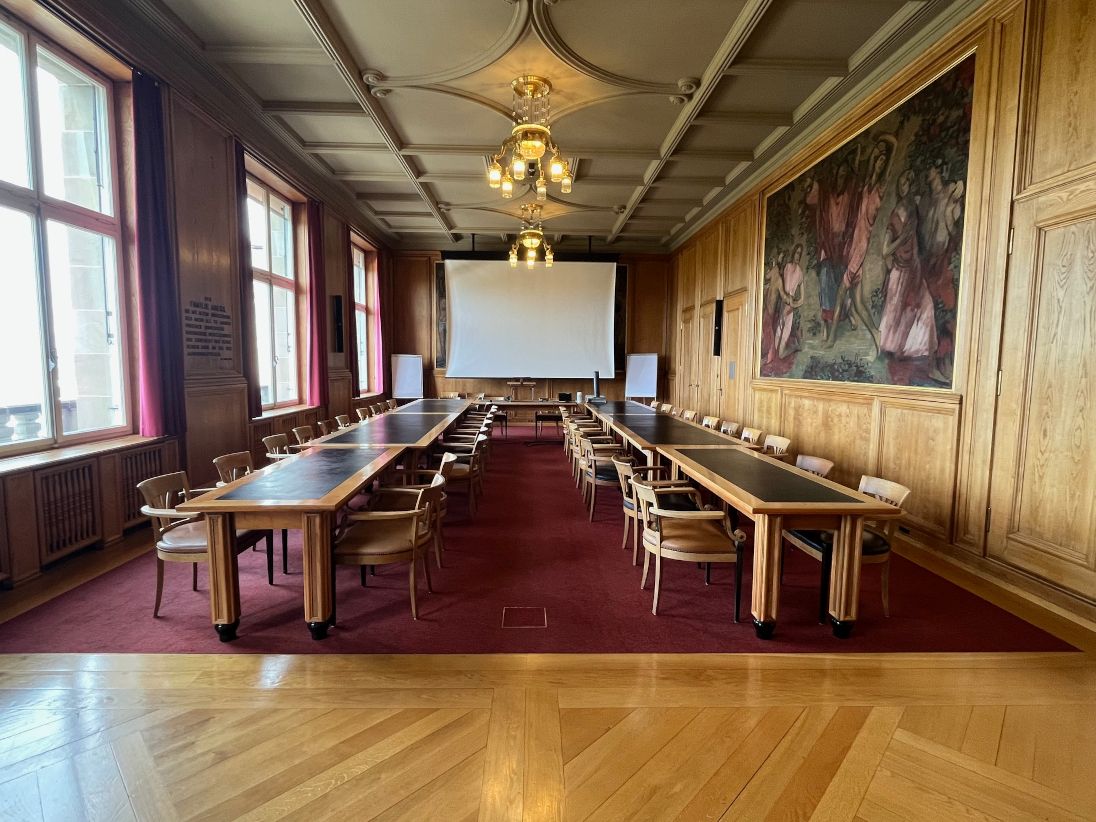 KOL E-13: Bild des Senatszimmers mit Sicht auf das Sprechpult. Die weisse Leinwand ist sichtbar. Die Eingänge befinden sich auf der rechten Seite.