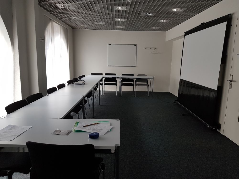 Seminarraum UNK-E-4: Der Seminarraum von der Eingangstüre aus gesehen. Die Wandtafel befindet sich hier rechts von der Türe. Die Tische sind U-förmig um den Raum platziert und können verschoben werden. Zudem ist die Bestuhlung nicht fix.