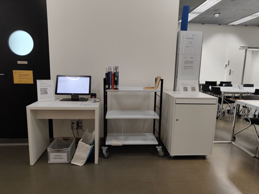 Arbeitsraum für Studierende KOL-H-303c: Links die Selbstausleihe (Hoehe der Tischflaeche 77cm), mittig der Bereitstellungswagen (Hoehe oberstes Tablar 97cm) und rechts der Rueckgabekasten (Hoehe Einwurf 87cm).