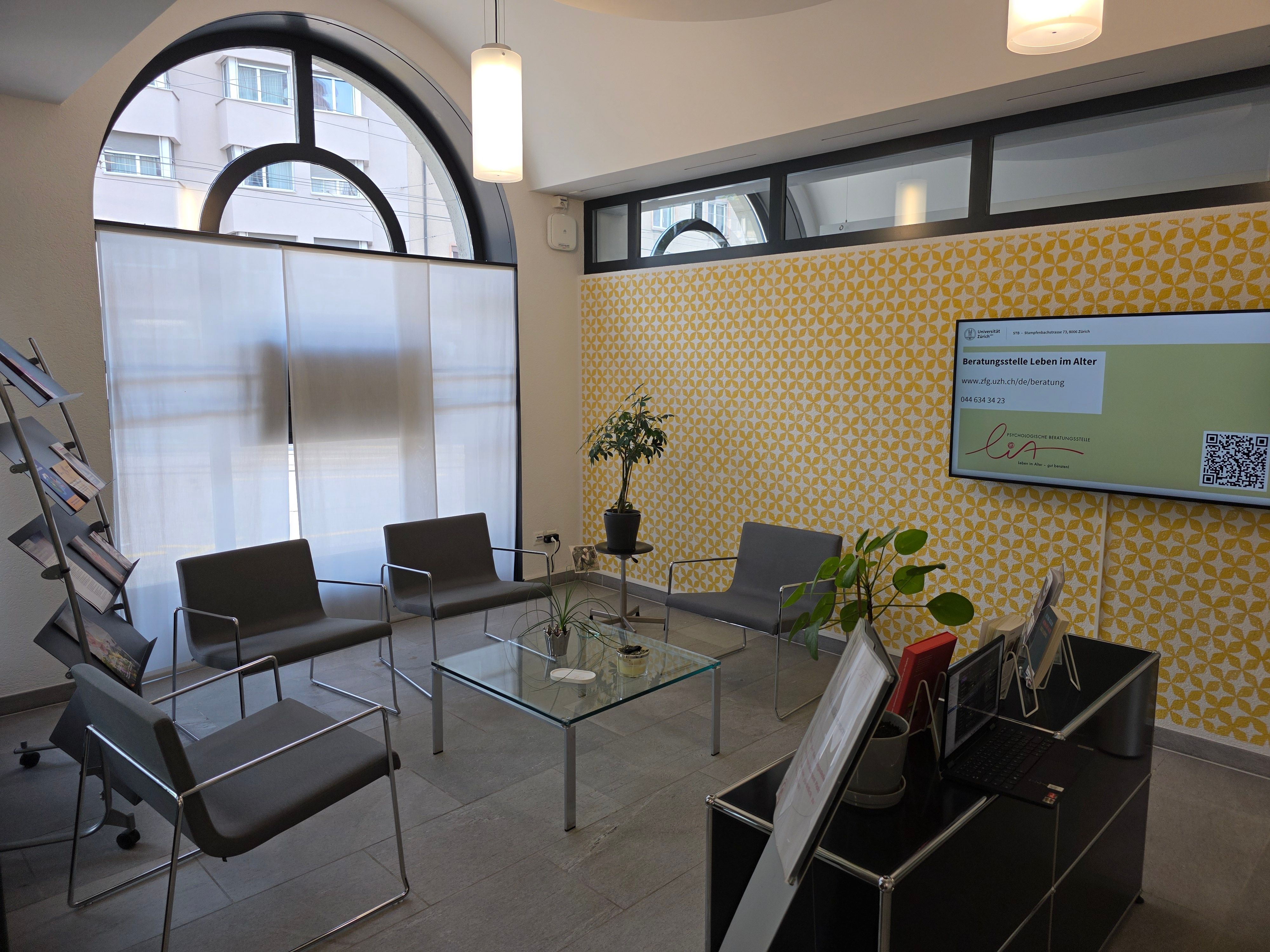 Foyer E-805: Wartebereich der Beratungsstelle im Alter. Vier graue Stühle stehen rund um einen kleinen Glastisch. Ein Regal mit diversen Broschüren steht daneben.