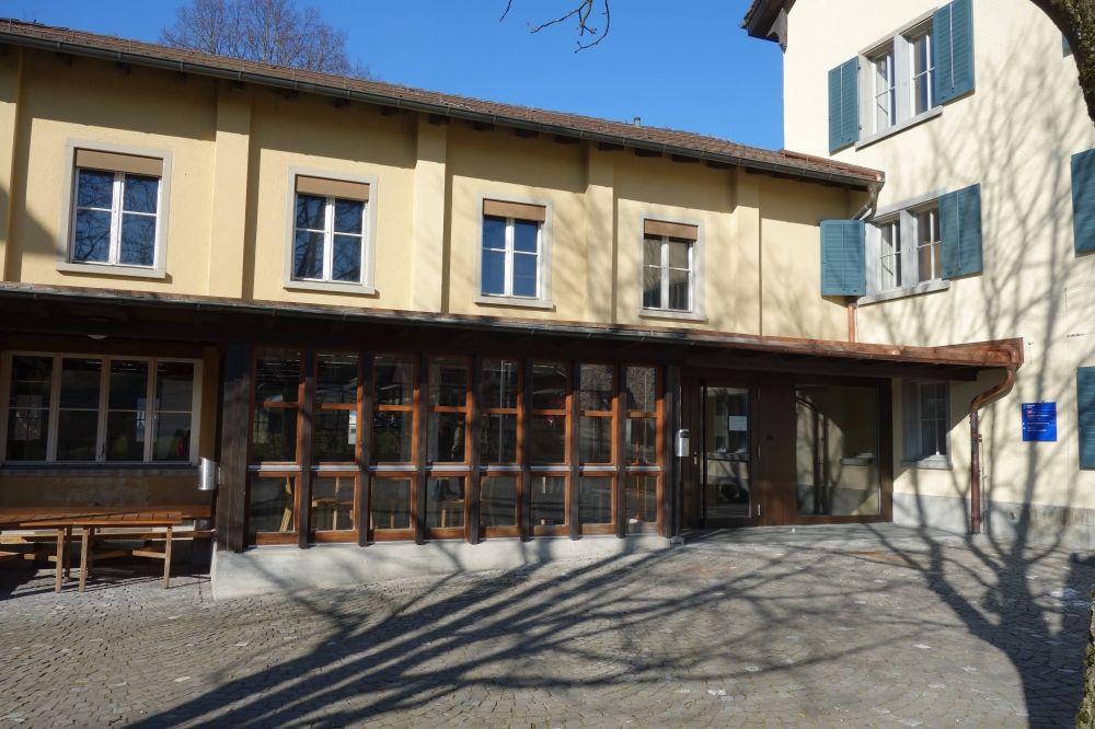 Gebäude Y63: Rechts der Haupteingang im Zwischentrakt des
                            Gebäudes. Es besteht eine leichte Steigung vor dem Eingang. Links sind
                            die überdachten Tische und Bänke zu sehen.