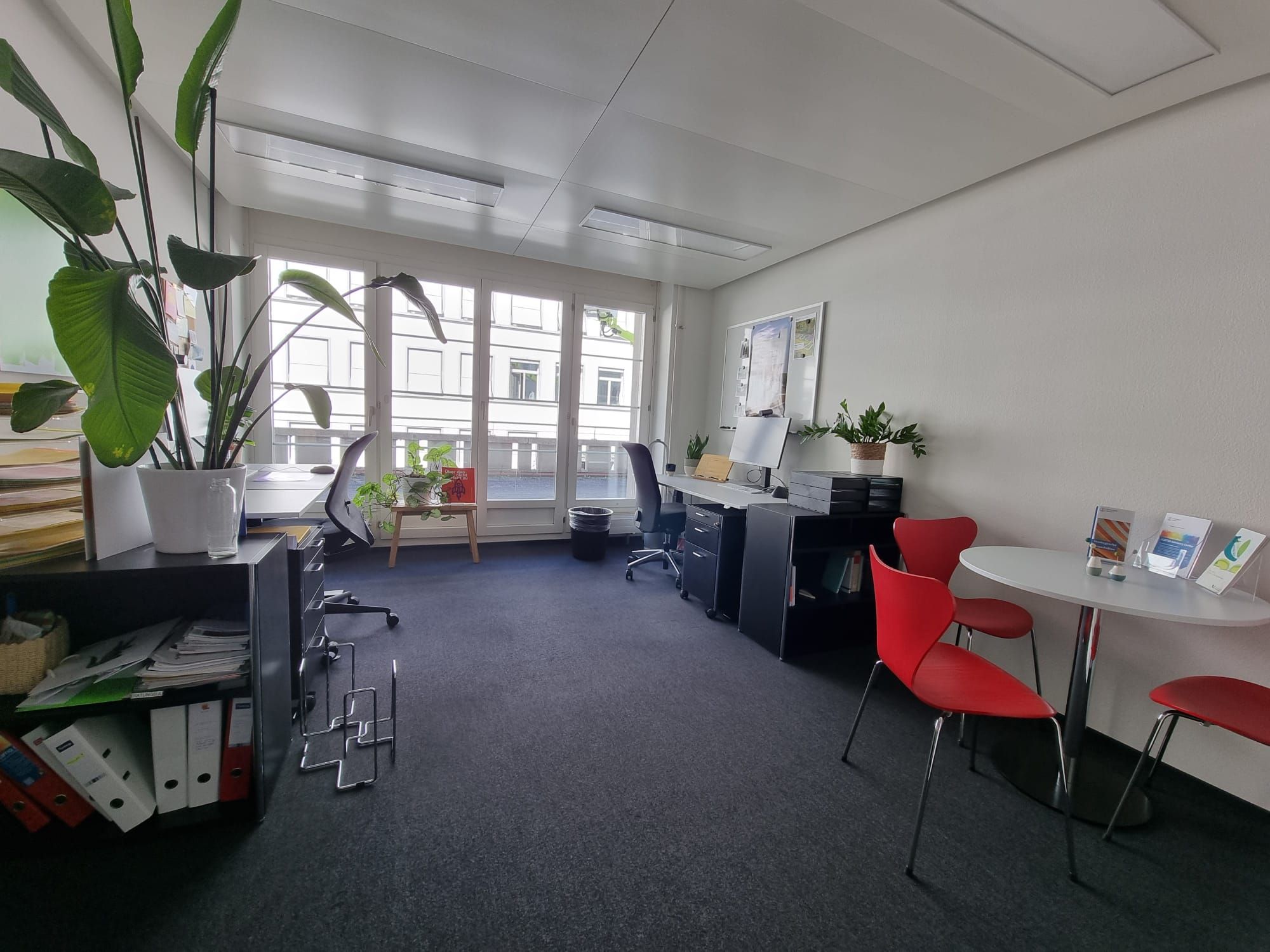 Da 015: Sicht von der Tür aus auf das Büro. Im Raum befinden sich zwei Büroplätze. Rechts vom Eingang hat es einen kleinen runden Tisch mit drei roten Stühlen rundherum. Die Wand gegenüber vom Eingang hat hohe Fenster und führt zur Terrasse.