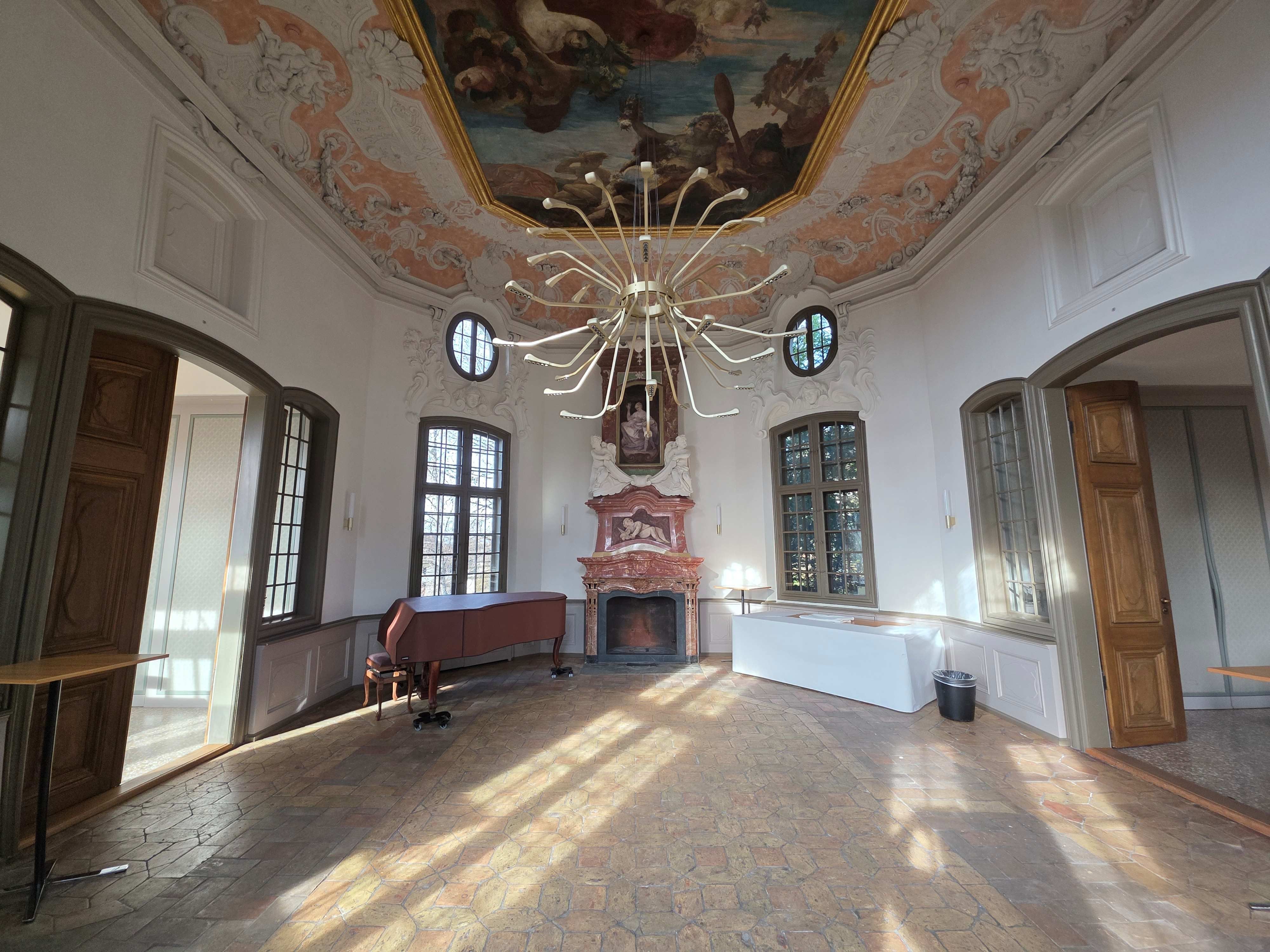 Eingangshalle E-01: Die achteckige Halle hat ein barockes Deckengemälde.  Auf der linken Seite hat es einen Flügel. Die Türen zu den zwei Sitzungszimmer befinden sich links und rechts vom Eingang.