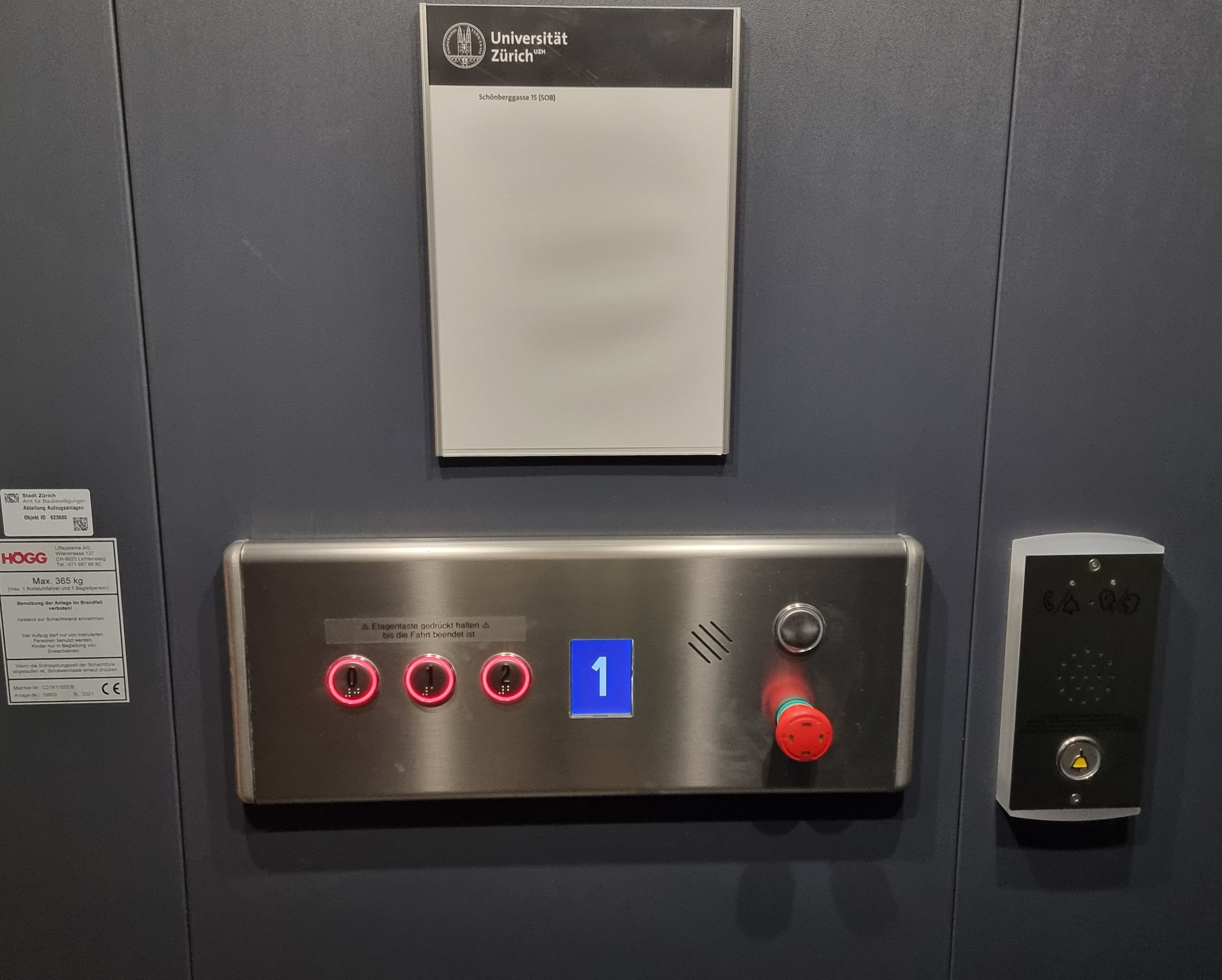 E-400J: Bild vom Bedienungsfeld im Inneren des Lifts. Es hat drei Tasten für die Stockwerke, 0, 1, und 2. Der Rand der Tasten leuchtet rot. Die Tasten sind beschriftet mit Relief-und Brailleschrift. Oberhalb der Tasten ist ein Kleber auf dem steht: "Etagentaste gedrückt halten bis die Fahrt beendet ist". Neben den Tasten ist ein rechteckiges Display. Auf dem Display sieht man die Zahl 1 in weisser Farbe mit einem blauem Hintergrund. Der Lift befindet sich auf dem 1. Stock.
Rechts neben dem Haupt-Bedienungsfeld befindet sich die Notfall-Sprechanlage. Darauf ist eine gelbe Klingeltaste abgebildet in Reliefschrift.