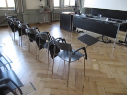 Hörsaal KAB-G-01: Am Stuhl montierte, ausklappbare Ablagefläche sowie Stromanschluss in der zweiten Reihe am Boden.