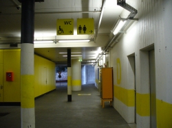 Rollstuhl-WC Y50-D-08A: Die erste Türe rechts am Bildrand ist der Zugang zur Rollstuhl-Toilette für Herren. Auf dem Bild ist der Raum vor den Aufzügen, dem Treppenhaus und dem Ticketautomaten des Stockwerks D.